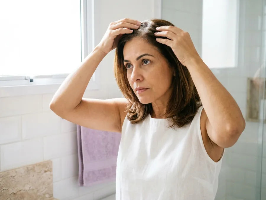 Mulher na frente do espelho abrindo a risca do cabelo com os dedos para olhar a raiz
